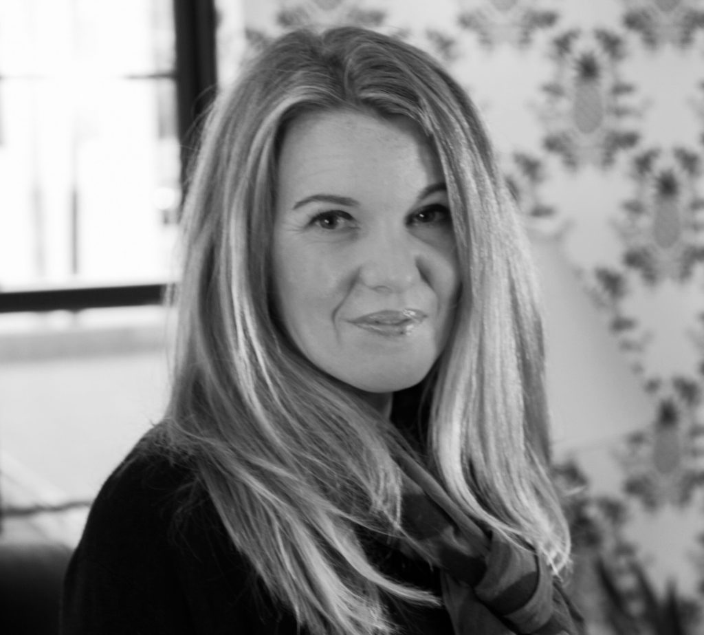 Professional portrait of Amanda J. Biedess, a woman with long, straight light-colored hair, wearing a dark top and looking directly at the camera with a subtle smile, softly lit indoors near a window with blurred foliage visible in the background, in black and white