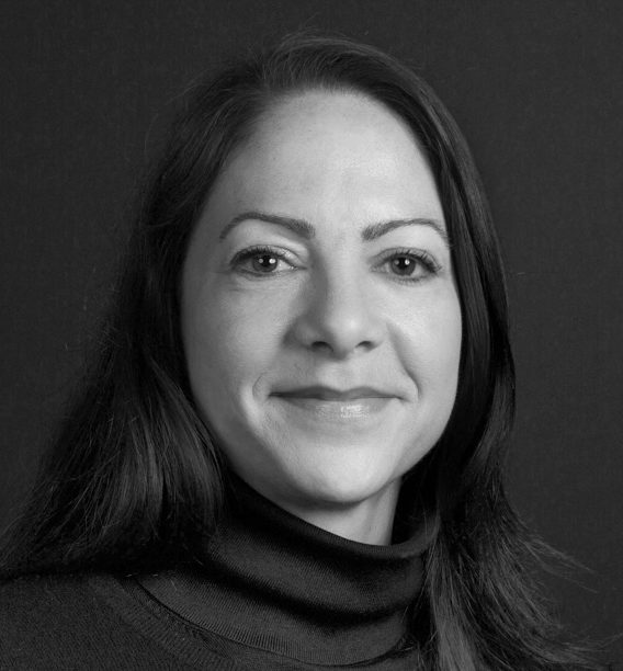 Professional headshot of Mary Kay Neuhaus, a woman with long dark hair, wearing a dark turtleneck sweater, smiling gently at the camera against a plain dark studio background, in black and white